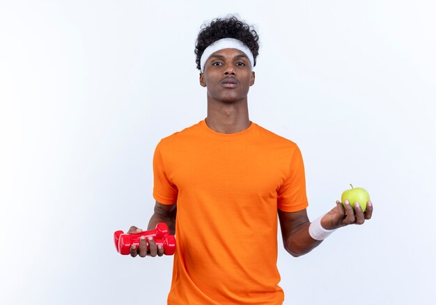 Patrząc na aparat fotograficzny młody afro-amerykański sportowy mężczyzna noszenie opaski i nadgarstka trzymając hantle z jabłkiem na białym tle