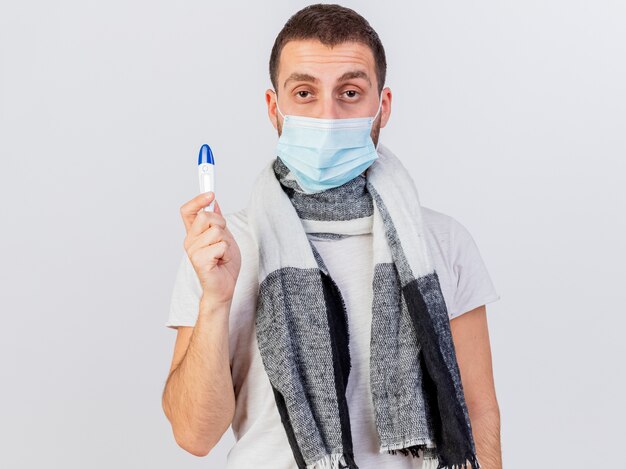 Patrząc na aparat młody chory człowiek ubrany w maskę medyczną i szalik trzyma termometr na białym tle