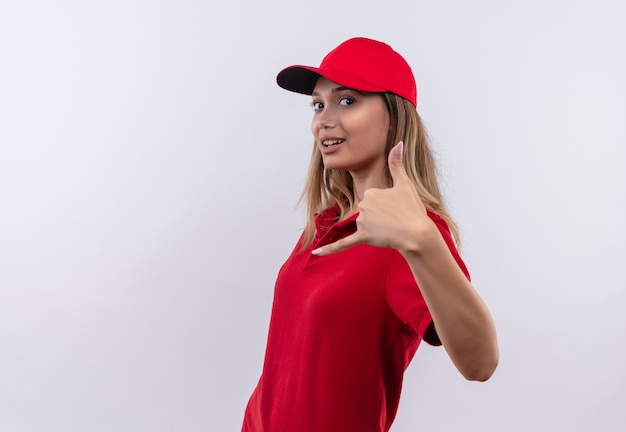 Patrząc na aparat zadowolony młoda dziewczyna dostawy ubrana w czerwony mundur i czapkę pokazując telefon gest na białym tle