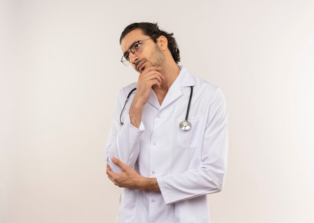 Patrząc na boczne myślenie młody lekarz mężczyzna z okularami optycznymi na sobie białą szatę ze stetoskopem, kładąc rękę na brodzie