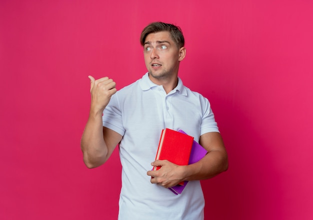 Patrząc na boczne myślenie młody przystojny student płci męskiej trzymający książki i punkty z boku na białym tle na różowej ścianie z miejsca na kopię