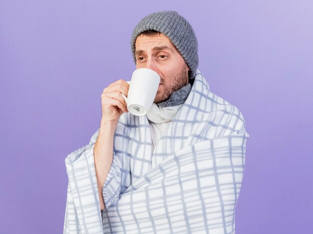 Patrząc na bok młody chory w czapce zimowej z szalikiem zawiniętym w kratę i pije herbatę na białym tle na fioletowym tle