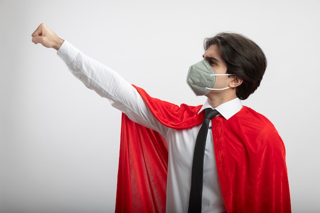 Bezpłatne zdjęcie patrząc na bok młody superbohater facet ubrany w krawat i maskę medyczną, podnosząc pięść na białym tle