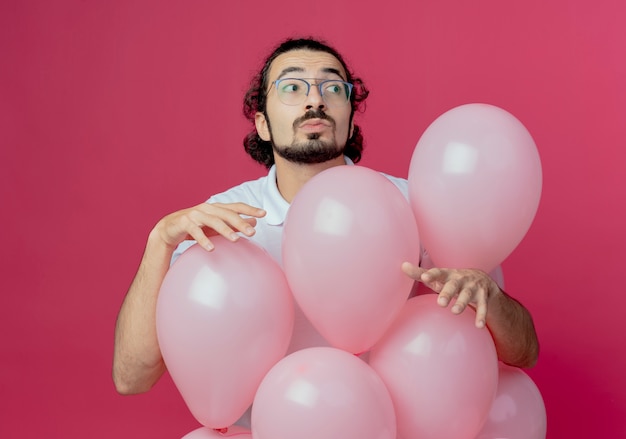 Patrząc na bok myślący przystojny mężczyzna w okularach stojący za balonami na białym tle na różowym tle