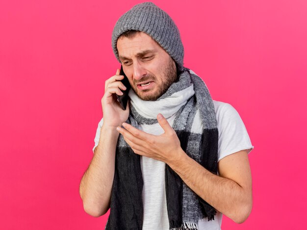 Patrząc na bok smutny młody chory w czapce zimowej z szalikiem mówi przez telefon na białym tle na różowym tle