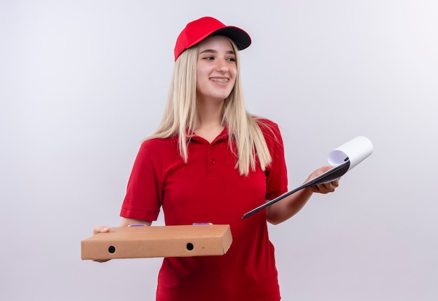 Bezpłatne zdjęcie patrząc na bok uśmiechnięta dostawa młoda kobieta ubrana w czerwoną koszulkę i czapkę, trzymając pudełko po pizzy i schowek na odosobnionej białej ścianie