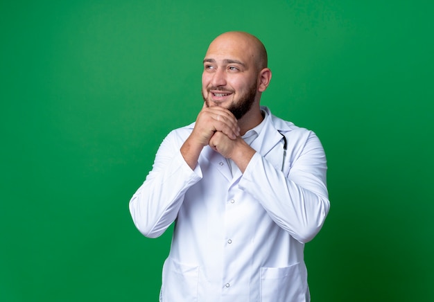 Patrząc na bok zadowolony młody lekarz mężczyzna ubrany w szlafrok i stetoskop kładąc ręce pod brodą na białym tle na zielonym tle