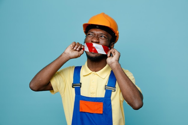 Patrząc na bok zapieczętowane usta taśmą młody afroamerykański budowniczy w mundurze na białym tle na niebieskim tle