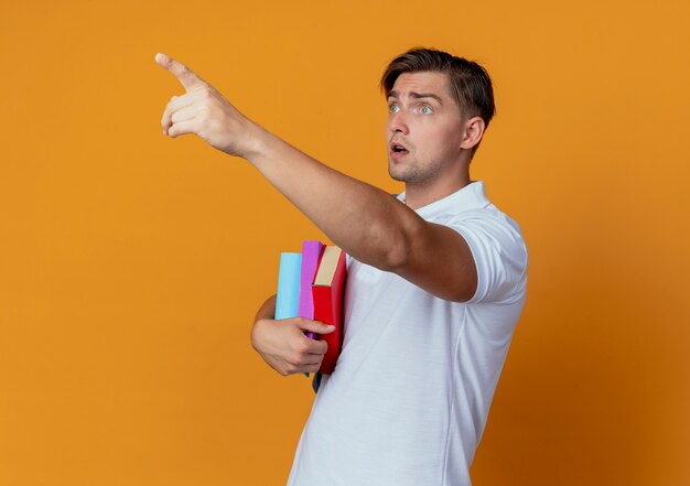 Patrząc na bok zaskoczony młody przystojny student płci męskiej ubrany w tylną torbę trzymając książki i wskazuje na bok na białym tle na pomarańczowym tle
