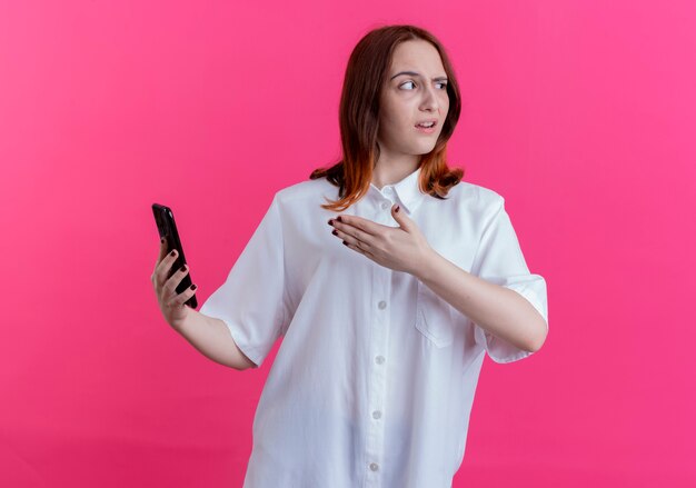Patrząc na boku niezadowolona młoda ruda dziewczyna trzyma i wskazuje ręką na telefon na białym tle na różowym tle