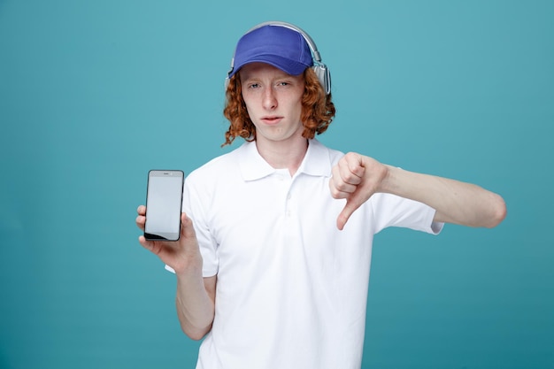 Patrząc na kamerę pokazującą kciuk w dół młody przystojny facet w czapce w słuchawkach, trzymający telefon na białym tle na niebieskim tle