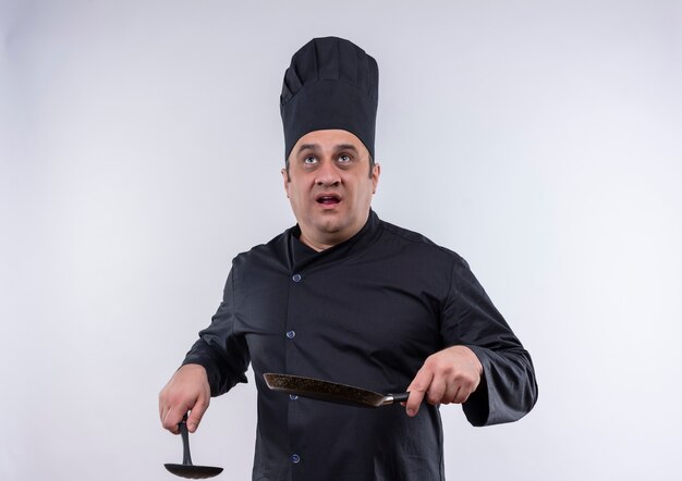 Patrząc na mężczyzna w średnim wieku kucharz w mundurze szefa kuchni, trzymając patelnię i łopatkę na odizolowanej białej ścianie