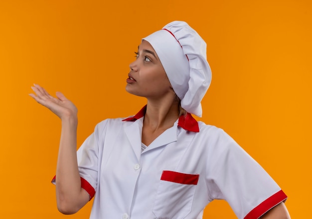 Patrząc na stronę młoda kobieta kucharz ubrana w mundur szefa kuchni wyciągając rękę na bok na odizolowanych pomarańczowej ścianie z miejsca na kopię