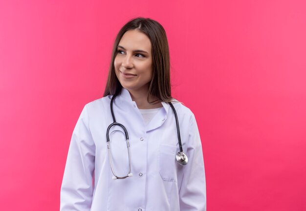 Patrząc na stronie młoda dziewczyna lekarza noszenie stetoskop medycznej sukni na na białym tle różowym