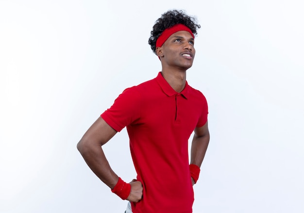 Patrząc na zadowolony młody afro-amerykański sportowy mężczyzna ubrany w opaskę i opaskę, kładąc ręce na biodrze na białym tle