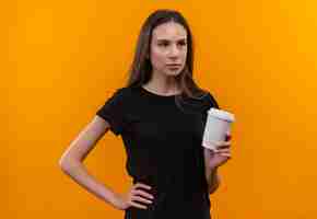 Bezpłatne zdjęcie patrząc z boku młoda dziewczyna kaukaska ubrana w czarną koszulkę trzymająca filiżankę kawy położyła dłoń na biodrze na odosobnionym pomarańczowym tle