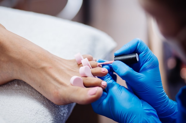 Pedicure w salonie domowym Pielęgnacja stóp i paznokci Proces profesjonalnego pedicure Mistrz w niebieskich rękawiczkach nakłada jasnoróżowy lakier hybrydowy