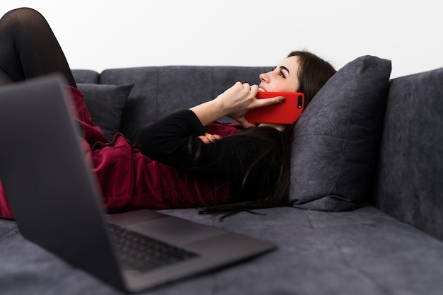 Pełna długość atrakcyjna kobieta, leżąc na kanapie i rozmawiając przez telefon komórkowy i laptop podczas pracy z domu.