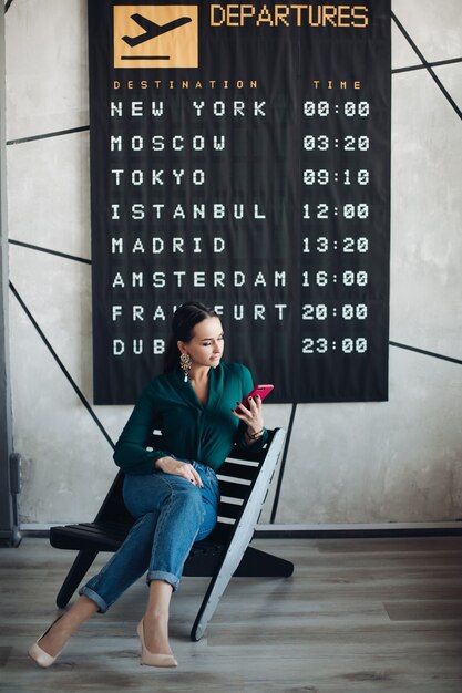 Pełna długość pień fotografia eleganckiej bizneswoman w smart casual przy użyciu telefonu komórkowego, czekając na jej lot przed plakatem odlotu.