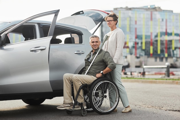Pełna długość portret dojrzałej pary z mężczyzną na wózku inwalidzkim samochodem na parkingu na zewnątrz miejsca kopiowania