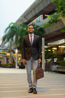 Pełna długość portret przystojnego młodego afrykańskiego biznesmena uśmiechającego się na zewnątrz w mieście