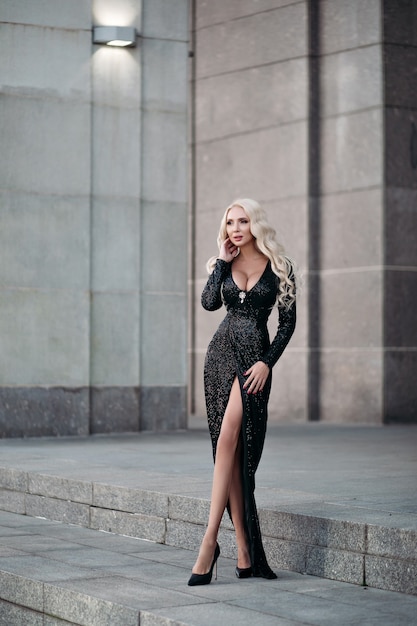 Pełna długość przepięknej szczupłej blondynki z długimi falującymi włosami i dużym biustem pozuje na ulicy w błyszczącej czarnej sukience i szpilkach.