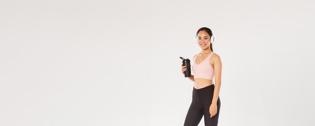 Pełna długość uśmiechniętej szczupłej i szczęśliwej dziewczyny fitness azjatycka kobieta trening athelte w siłowni z słuchawkami