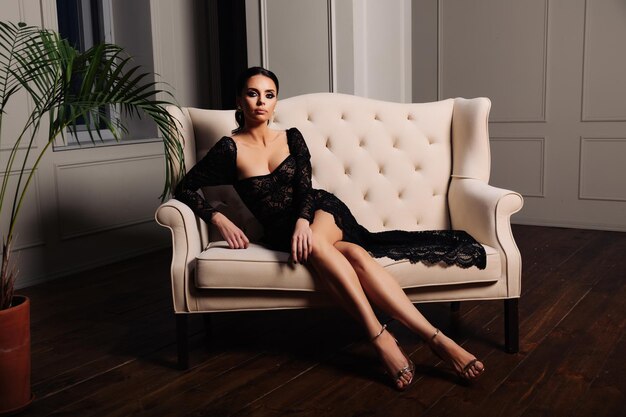 Pełna wdzięku opalona elegancka młoda kobieta w czarnej sukni wieczorowej siedząca na eleganckiej luksusowej sofie w ...