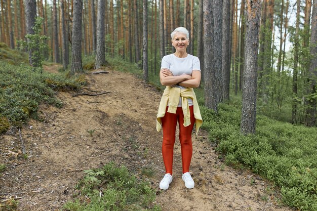 Pełne ujęcie aktywnej, dojrzałej kobiety o blond włosach i wysportowanej sylwetce stojącej na szlaku w lesie w sportowym stroju, odpoczywającej podczas ćwiczeń z założonymi rękami. Ludzie, aktywność i wiek