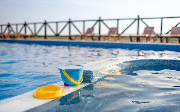 Pełne wiadro z wodą dla dzieci, żółte sitko i kilka innych zabawek plażowych znajdują się na łodzi z niebieskiego basenu z czystą przezroczystą wodą na tle ciepłego słonecznego letniego zachodu słońca