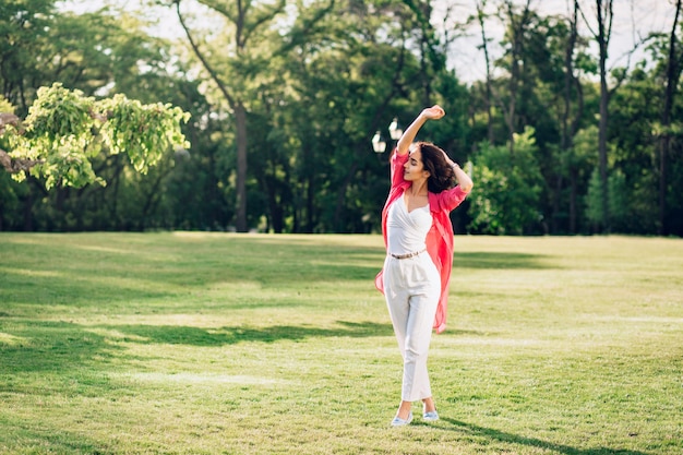 Pełne zdjęcie uroczej brunetki dziewczyny spacerującej w letnim parku. Nosi białe ubranie i długą różową koszulę. Ona się bawi.