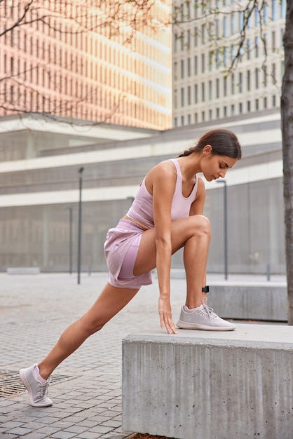 Pełnej długości ujęcie aktywnej muskularnej kobiety opiera się na betonowym kamieniu, ubranej w odzież sportową, rozgrzewa się przed treningiem pozuje na zewnątrz na tle miejskim Wysportowana modelka przygotowuje się do biegu