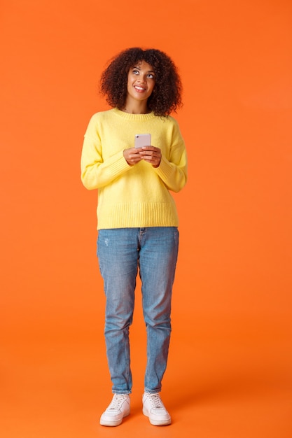 Pełnometrażowe pionowe ujęcie marzycielskiej uroczej afroamerykańskiej kobiety zastanawiającej się, co napisać, trzymając smartfon myślący, patrząc w górę i uśmiechając się, wyobrażając sobie rzeczy, stojąc radośnie na pomarańczowej ścianie.