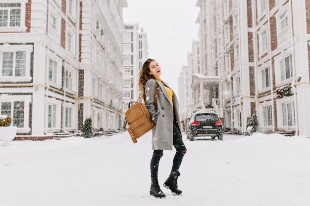 Pełnometrażowy portret europejki w eleganckim płaszczu w śnieżną pogodę. wesoła młoda kobieta ze stylowym plecakiem stojącym przy głównej ulicy miasta w zimowy dzień.