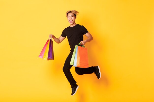 Pełnometrażowy szczęśliwy atrakcyjny Azjata skaczący ze szczęścia i niosący torby na zakupy, stojący na żółtej ścianie.