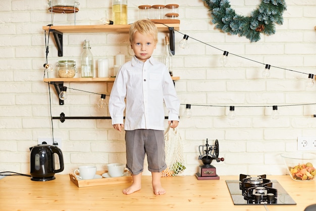 Pełnowymiarowy obraz uroczego chłopca z blond włosami stojącego boso na drewnianym stole w stylowym skandynawskim wnętrzu kuchni ze świątecznym wieńcem, źle się zachowującego, gdy nikt go nie widzi