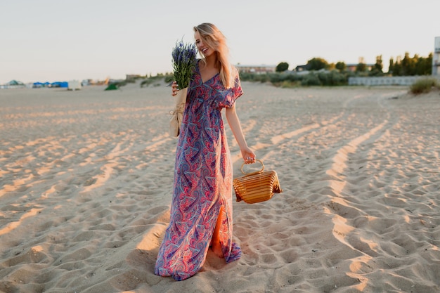 Pełnowymiarowy wizerunek ładnej blond kobiety z bukietem lawendy spacerującej po plaży. Kolory zachodu słońca.