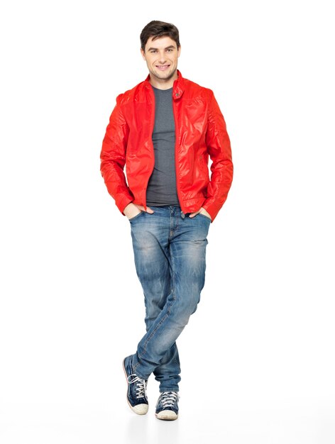 Pełny portret uśmiechnięty szczęśliwy przystojny mężczyzna w czerwonej kurtce, dżinsach i butach gimnastycznych. Piękny facet stojący na białym tle