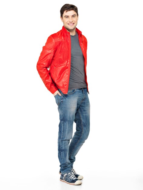 Pełny portret uśmiechnięty szczęśliwy przystojny mężczyzna w czerwonej kurtce, niebieskich dżinsach i butach gimnastycznych. Piękny facet stojący na białym tle