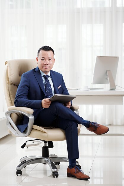 Pełny strzał udanego azjatyckiego dyrektora wykonawczego, siedzący ze skrzyżowanymi nogami na swoim luksusowym krześle szefa w lekkim przestronnym biurze