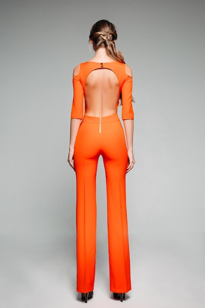 Bezpłatne zdjęcie pełny widok z tyłu modnego modelu w swobodnym jasnopomarańczowym kombinezonie z odkrytymi plecami i wysokimi obcasami na szarym tle