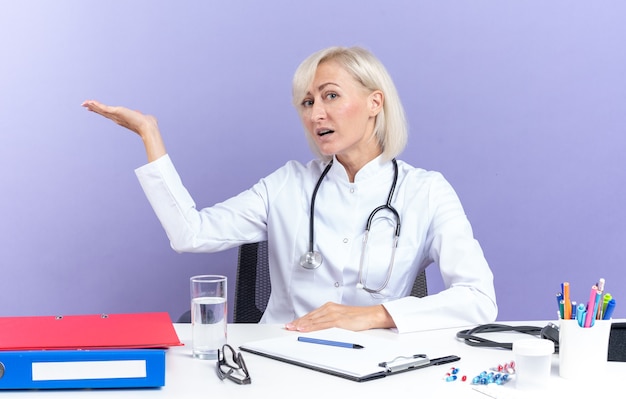 Bezpłatne zdjęcie pewna dorosła lekarka w szacie medycznej ze stetoskopem siedząca przy biurku z narzędziami biurowymi trzymająca rękę otwartą odizolowaną na fioletowej ścianie z miejscem na kopię