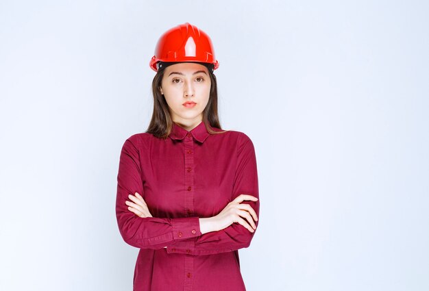 Pewna kobieta architekt w czerwonym kasku twardym stojąc i pozowanie na białej ścianie.