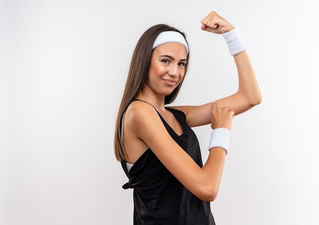 Pewna ładna sportowa dziewczyna nosząca opaskę i nadgarstek, gestykulując mocno i dotykając jej ramienia na białym tle na białej ścianie z miejscem na kopię