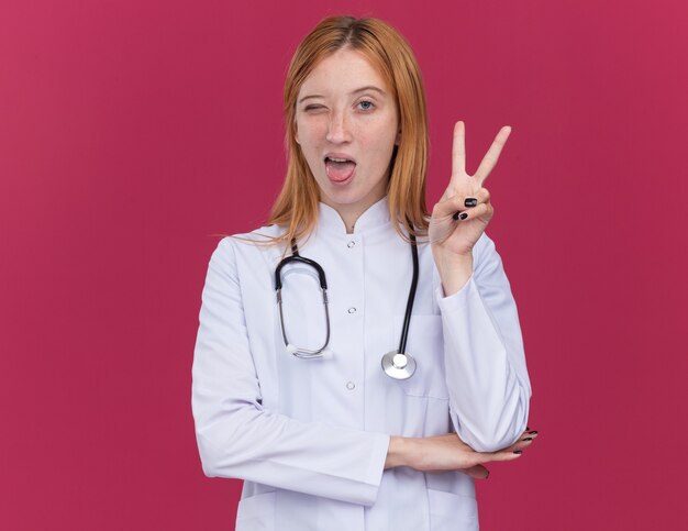 Pewna młoda kobieta imbir lekarz ubrany w szatę medyczną i stetoskop, patrząc na przód mrugając pokazując język robi znak pokoju odizolowany na szkarłatnej ścianie z kopią przestrzeni