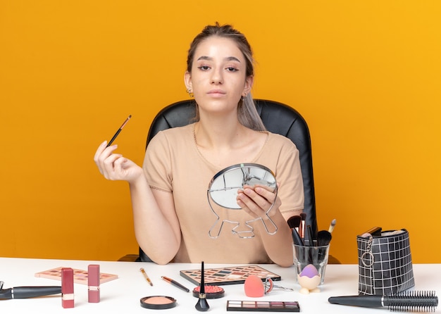 Bezpłatne zdjęcie pewna młoda piękna dziewczyna siedzi przy stole z narzędziami do makijażu, trzymając pędzel do makijażu z lustrem na białym tle na pomarańczowej ścianie