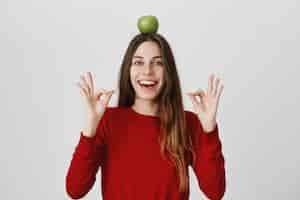 Bezpłatne zdjęcie pewna siebie i podekscytowana, uśmiechnięta śliczna kobieta pokazująca znak dobra i trzymająca zielone jabłko na głowie