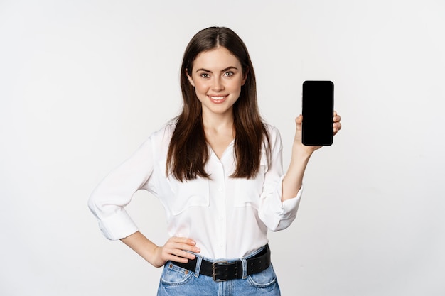 Pewna siebie kobieta w korporacyjnych ubraniach, pokazująca ekran smartfona, mobilny interfejs aplikacji, stojąca na białym tle.