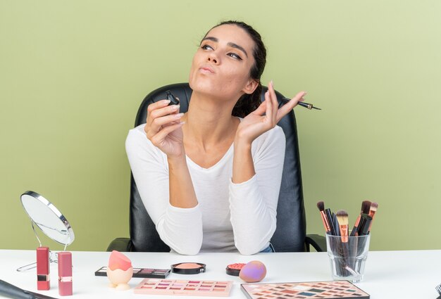 Pewna siebie, ładna kaukaska kobieta siedzi przy stole z narzędziami do makijażu, trzymając eyeliner i patrząc na bok