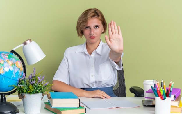 Pewna siebie młoda blond studentka siedząca przy biurku z szkolnymi narzędziami, wykonująca gest zatrzymania